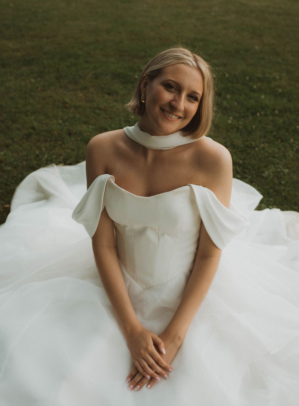 A smiling bride in an elegant off-shoulder white ball gown sits on green grass, wearing a choker necklace.