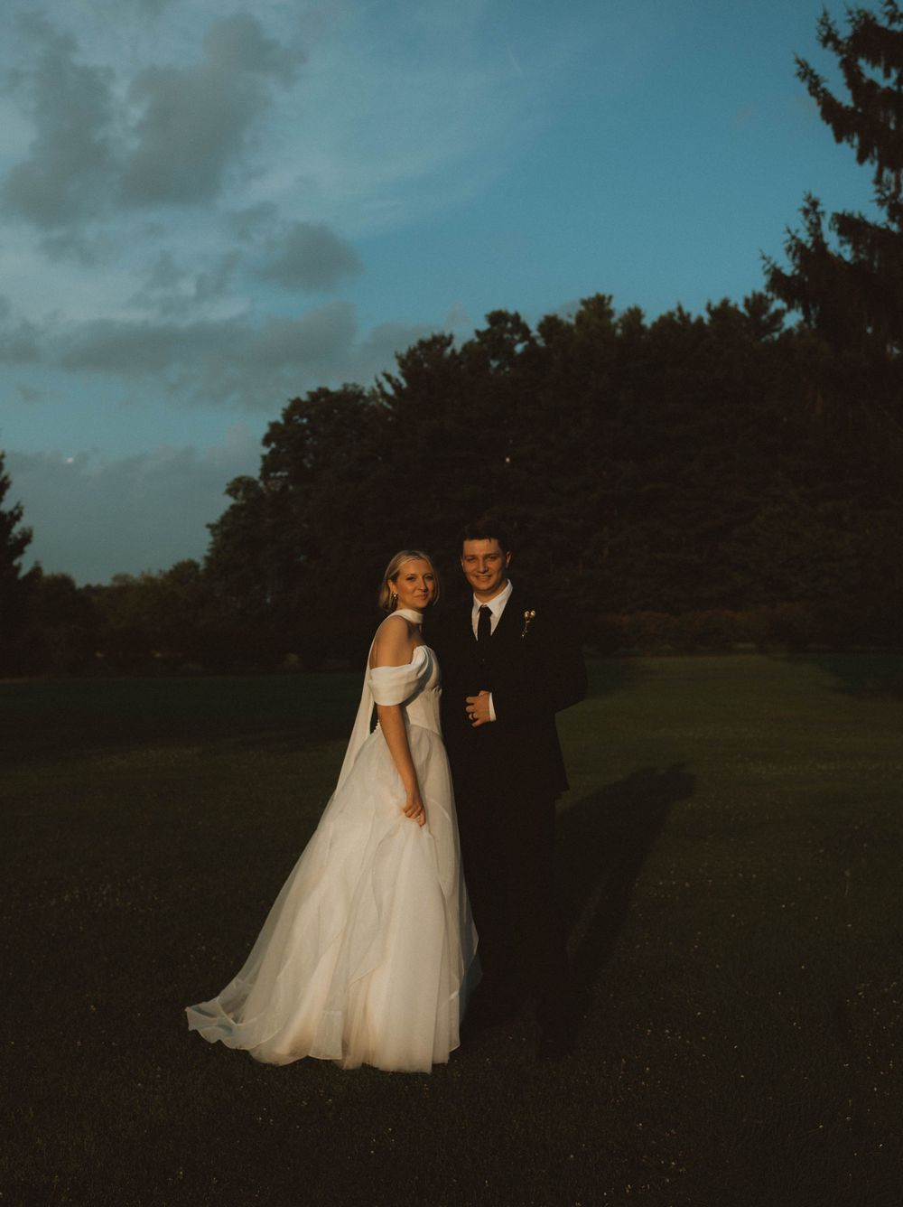 Bride in off-shoulder gown and groom in black suit pose outdoors at dusk on lush green lawn.