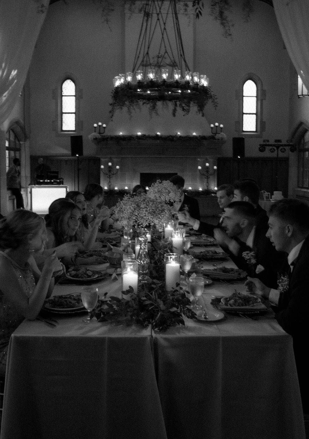 Black and white wedding reception dinner with guests seated at candlelit table beneath floral chandelier.