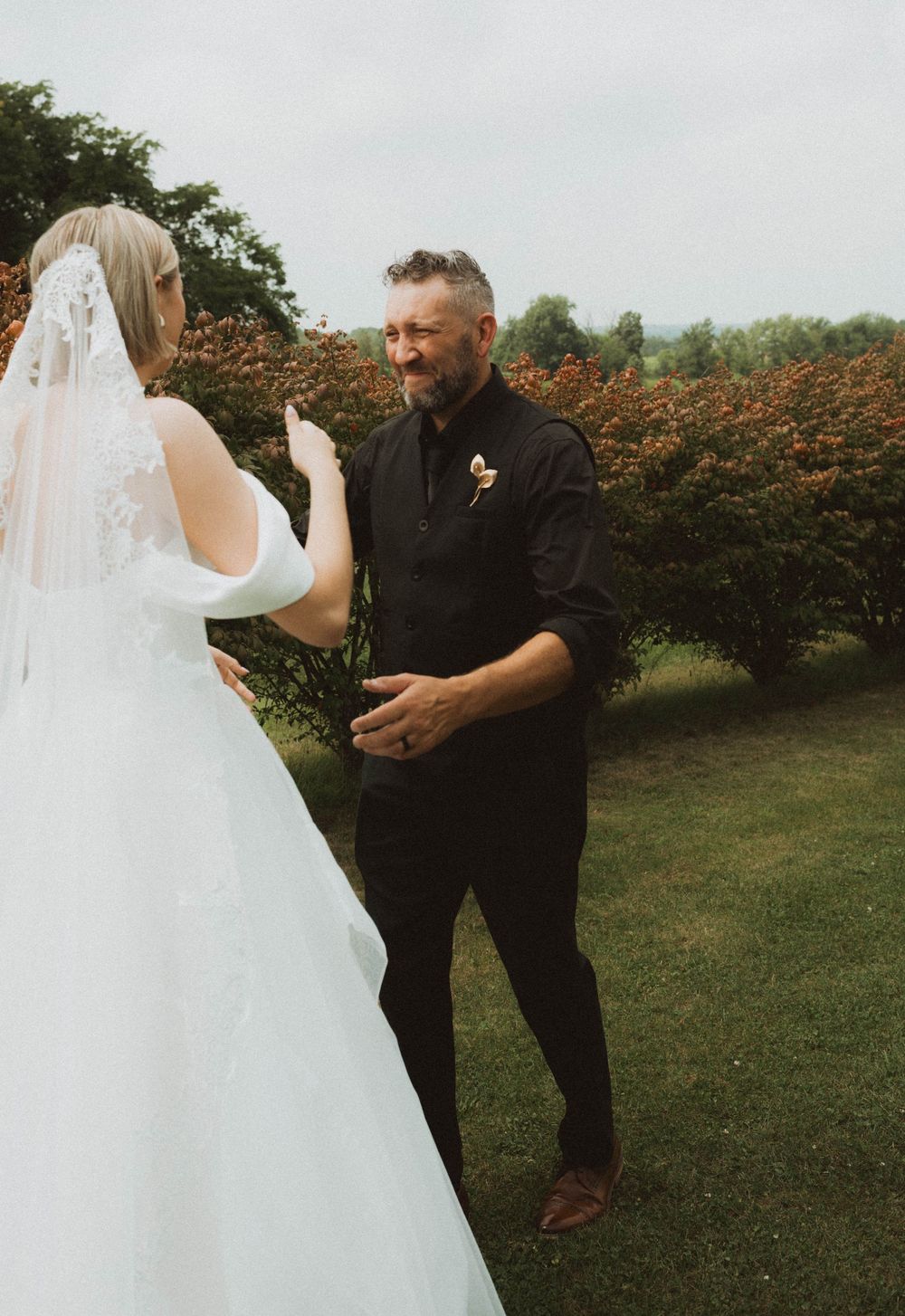 A beaming groom in black suit sees his bride for the first time at an outdoor wedding first look.
