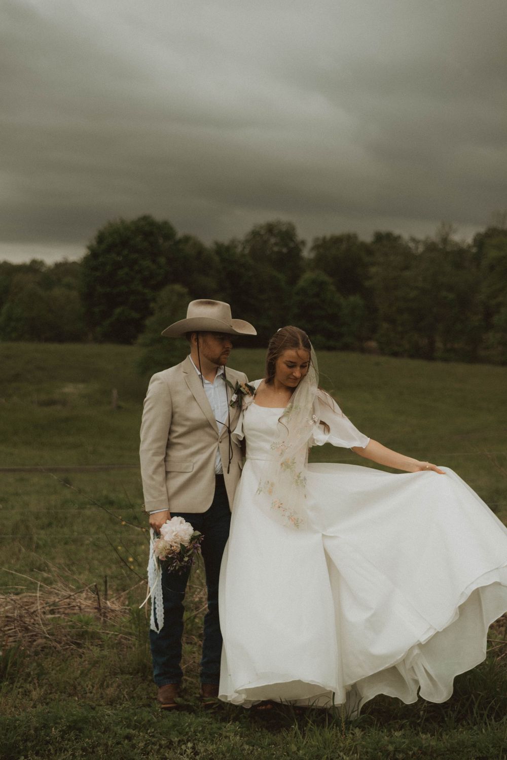 Bride in white dress and groom in tan cowboy hat pose together in a green field under dramatic stormy skies.