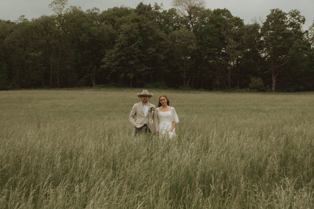 Couple walking hand in hand through tall green meadow grass with lush forest backdrop on overcast day.