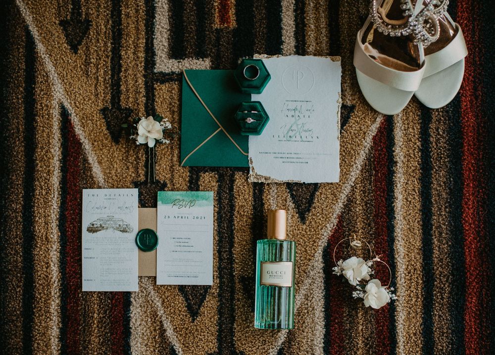 Elegant green and white wedding stationery suite with wax seals, perfume bottle, and bridal accessories on a patterned rug.