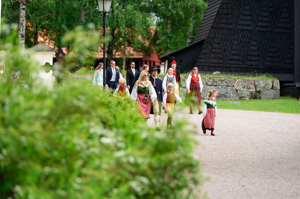 Group in traditional Scandinavian folk costumes walking along a gravel path near historic wooden buildings.