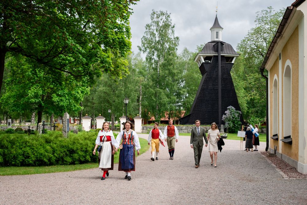 People in traditional Scandinavian folk costumes walking along a path near a historic wooden bell tower.