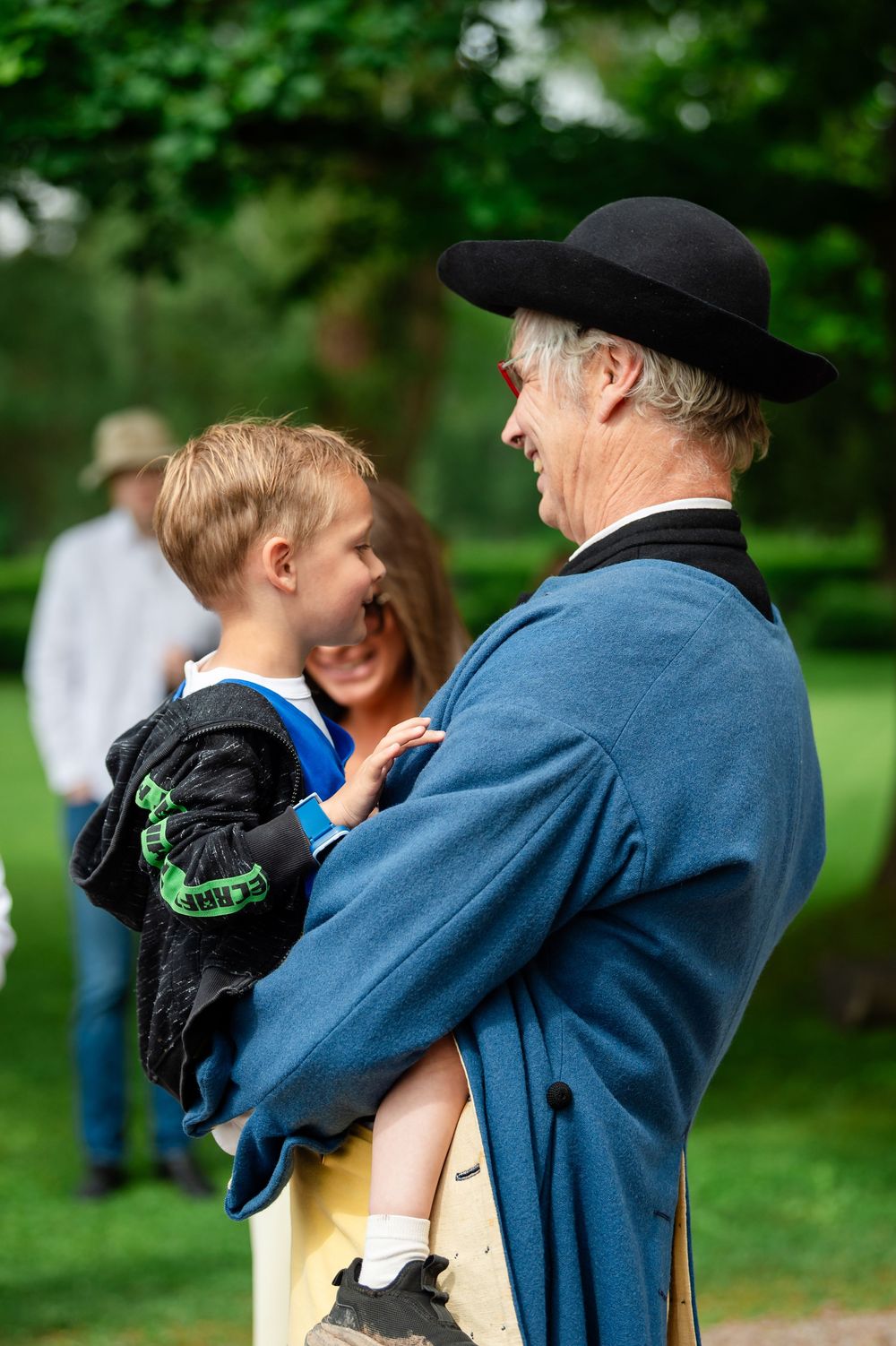 An elderly man in a blue coat and black hat lovingly holds a young blonde boy outdoors.