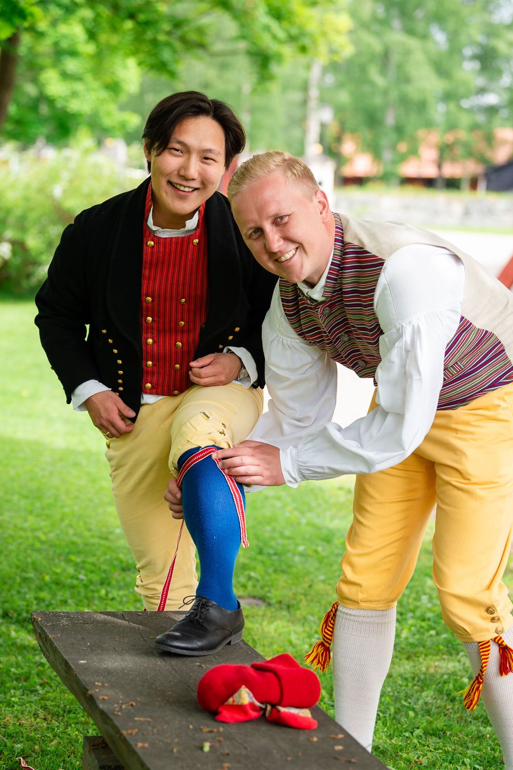 Two men in colorful traditional Scandinavian folk costumes smiling outdoors while adjusting their festive attire.