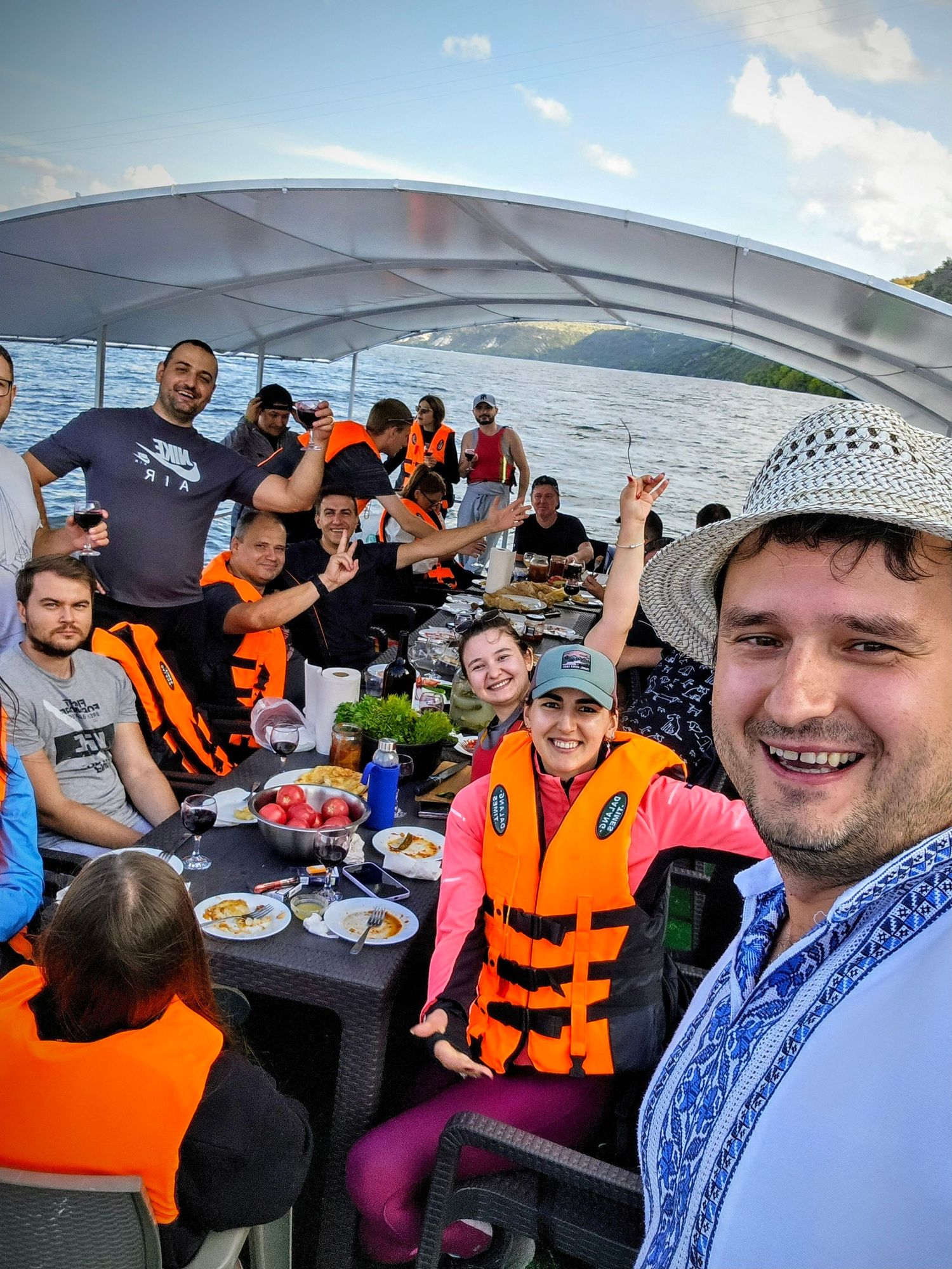 Happy group of tourists wearing orange life jackets dining on a boat on a scenic lake at sunset.