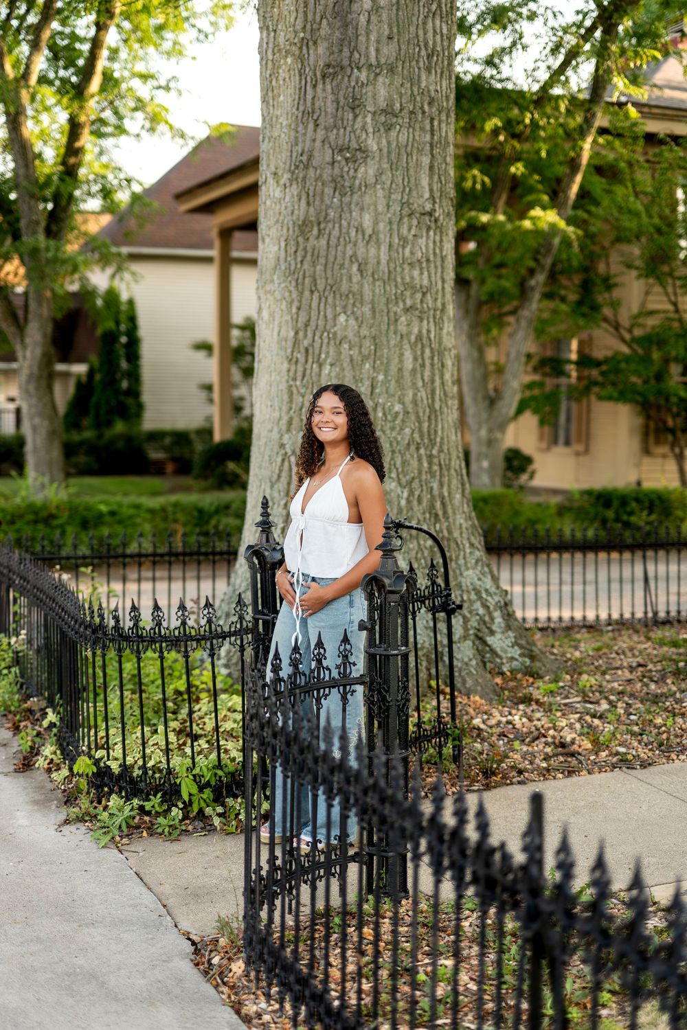 Smiling young woman in white top and jeans posing by iron gate near large tree.