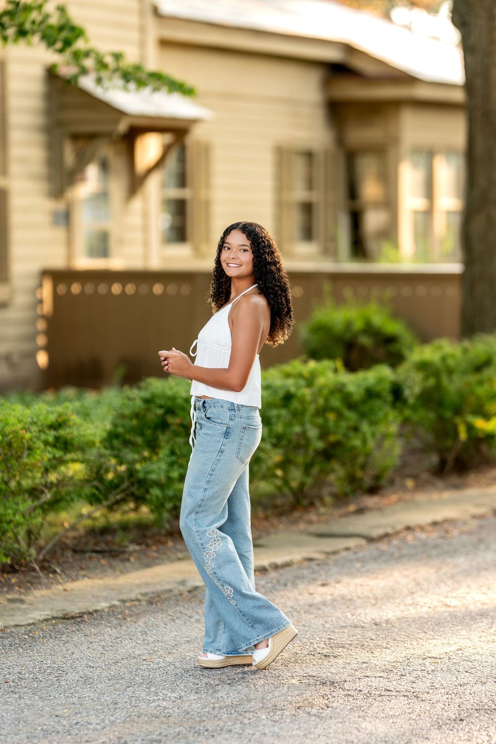 Young woman with curly hair in white top and wide-leg jeans walking on a sunlit residential driveway.