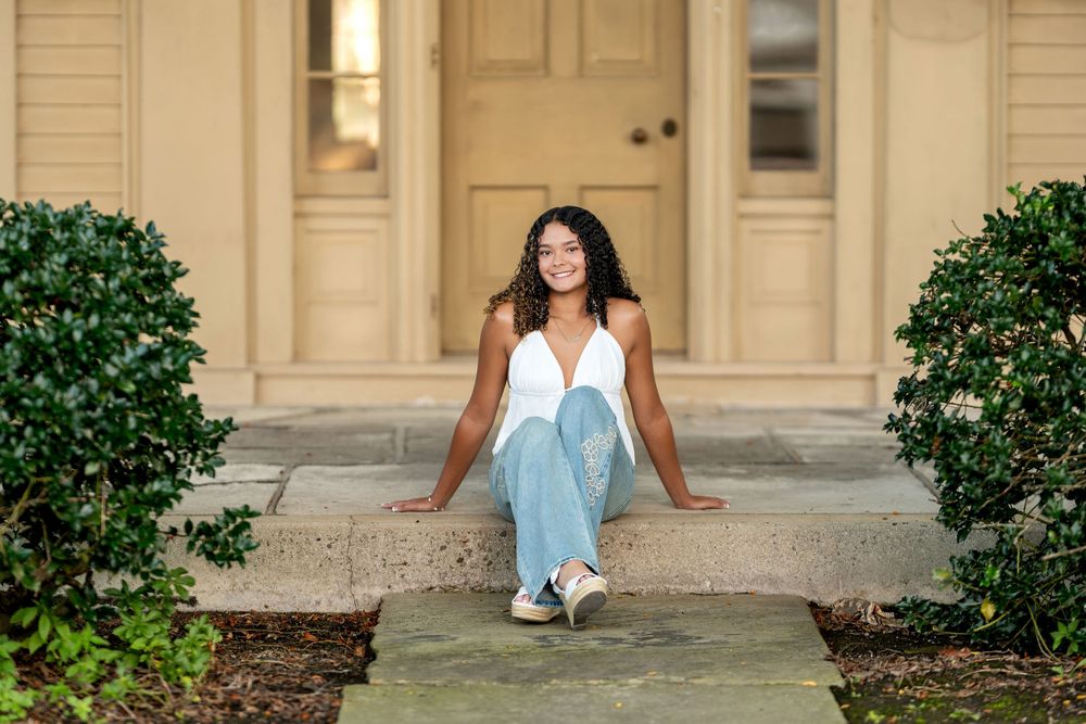 Young woman in white top and jeans sitting on steps in front of a beige house entrance.