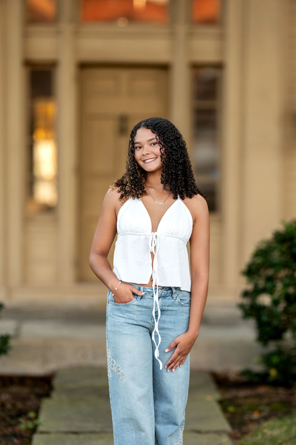 Smiling young woman in white crop top and jeans posing in front of elegant building entrance.
