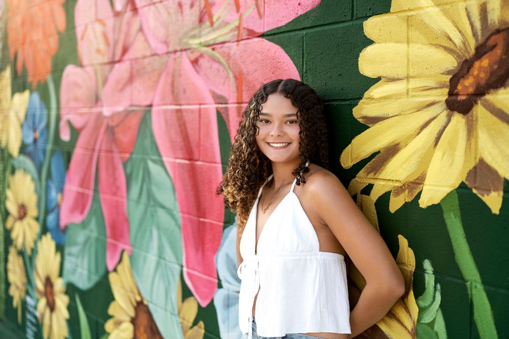 Smiling young woman in white top posing in front of a colorful floral mural outdoors.