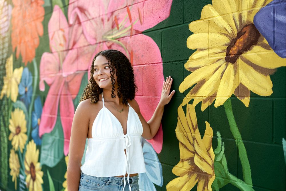 Smiling young woman in white top posing in front of colorful floral mural on brick wall.