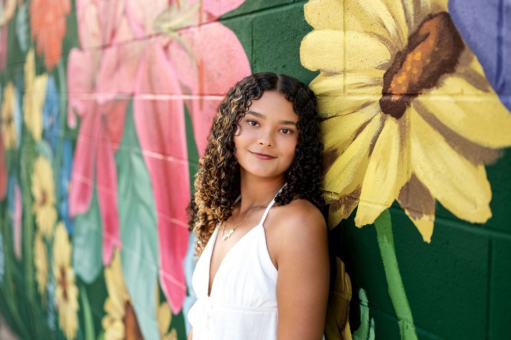 Young woman with curly hair leaning against vibrant sunflower painted brick mural.