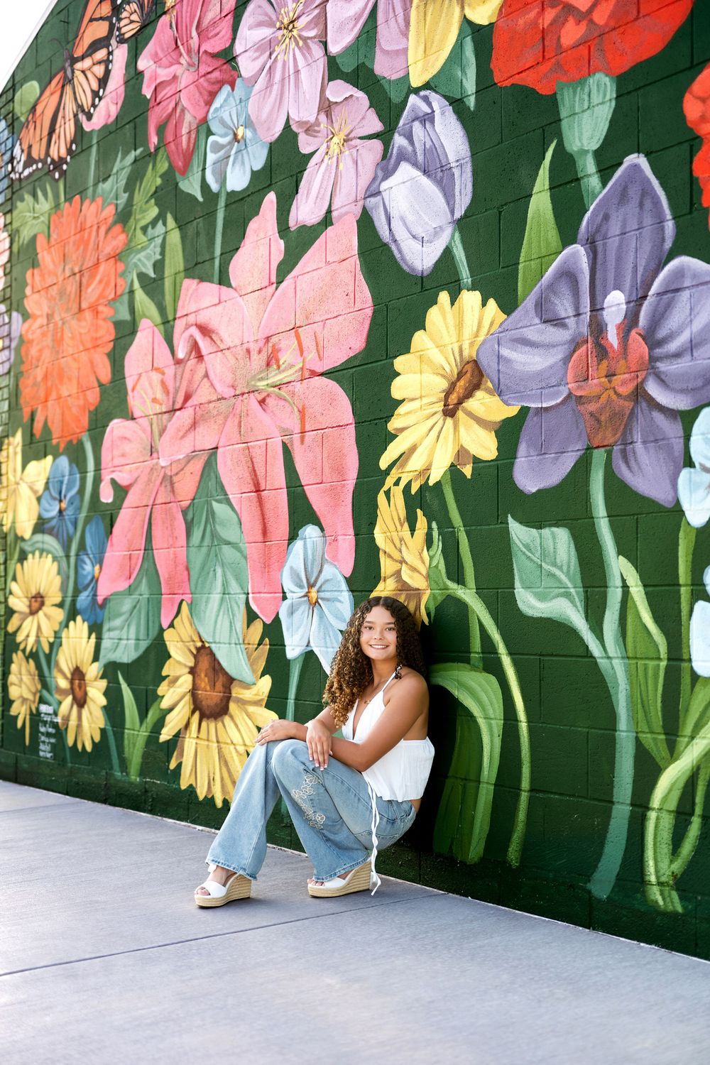 Young woman sitting against colorful floral mural on brick wall.