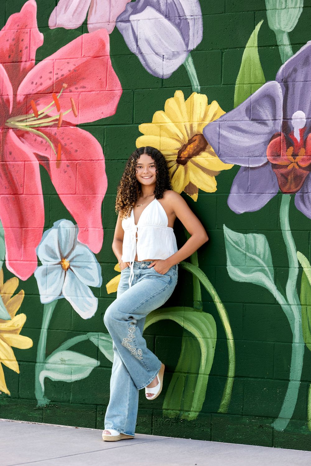 Young woman in white top and jeans leaning against vibrant floral mural on green brick wall.