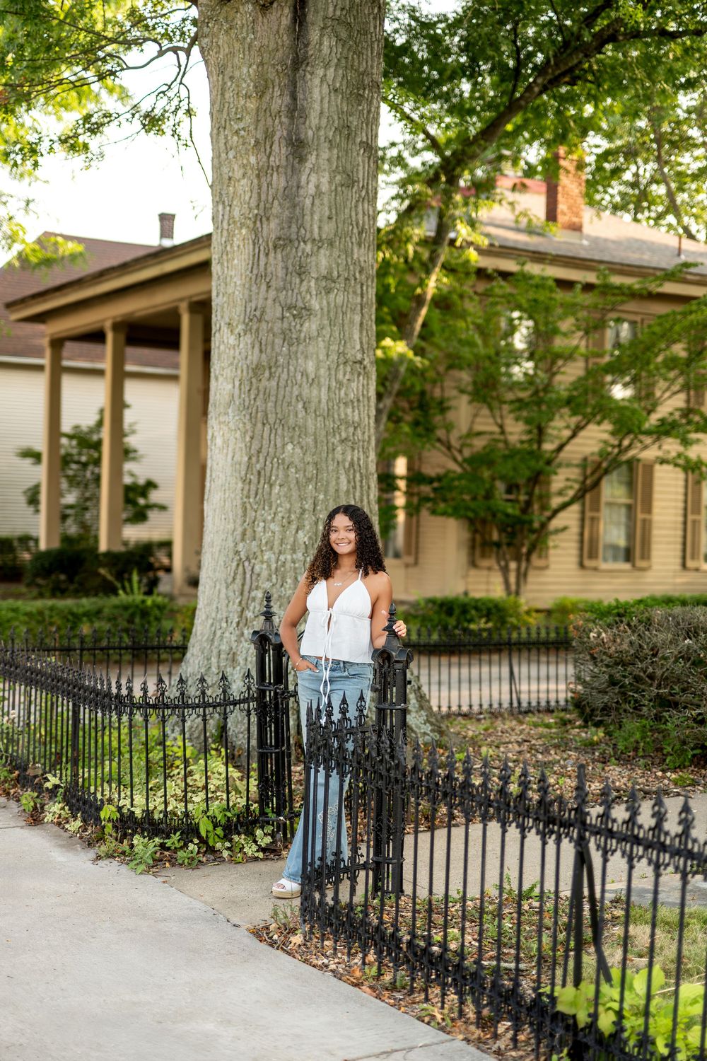 Young woman in white top and jeans leaning on iron fence by historic home.