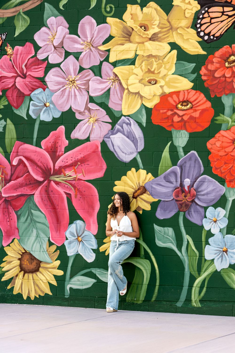 Young woman in white top and jeans posing in front of a vibrant colorful floral street mural.
