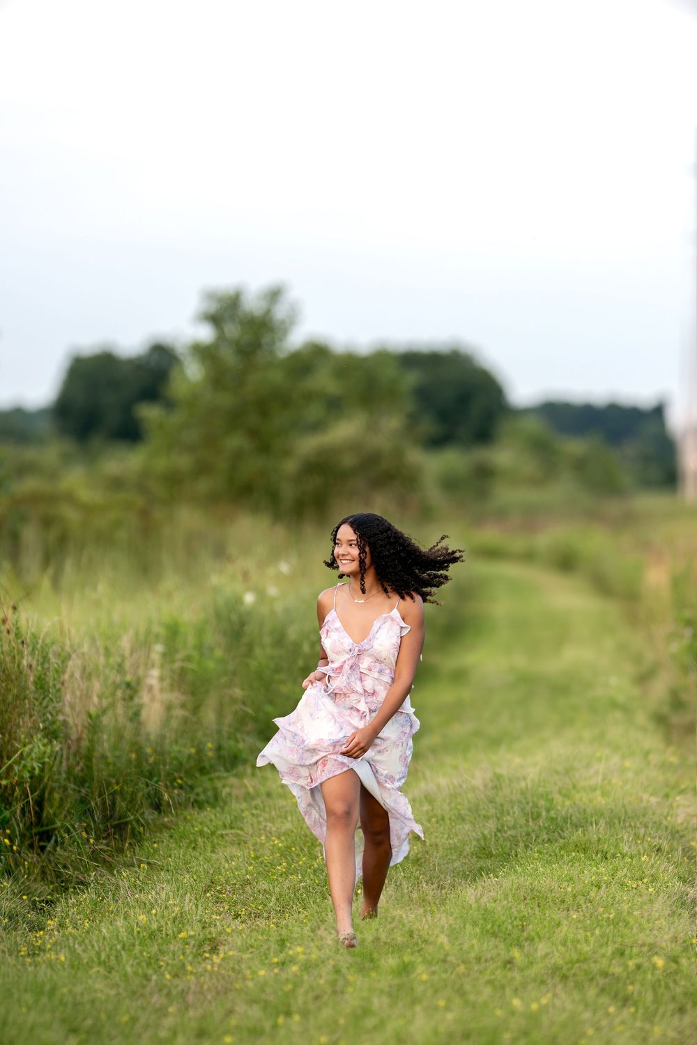 Young woman in floral dress running barefoot through green meadow path with hair flowing in wind.