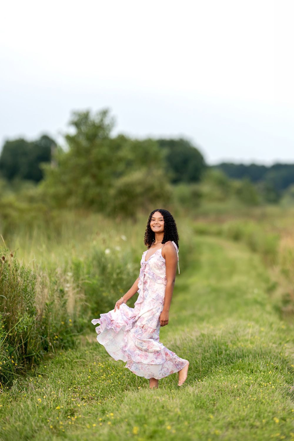 Young woman in floral dress twirling in green meadow on overcast day.