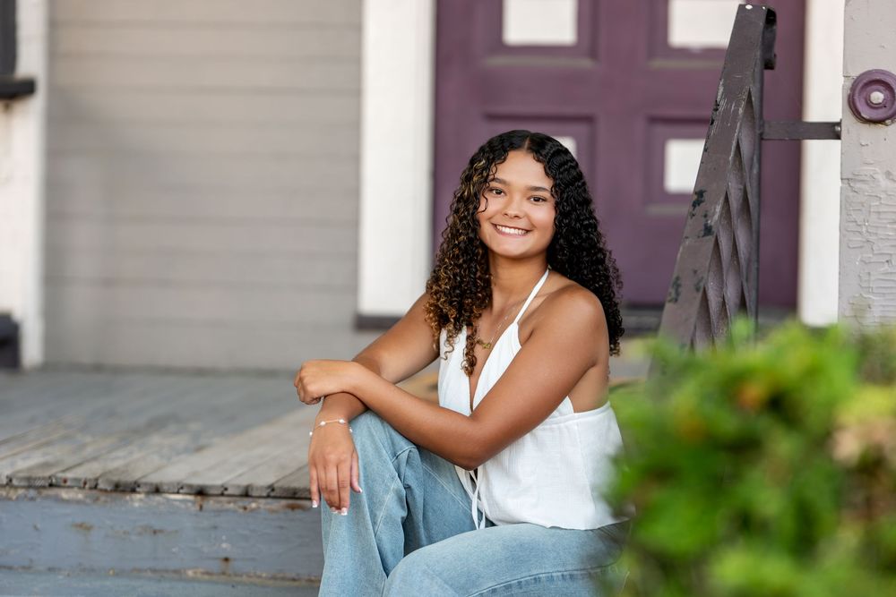Young woman with curly hair smiling on porch steps, wearing white tank top and jeans.