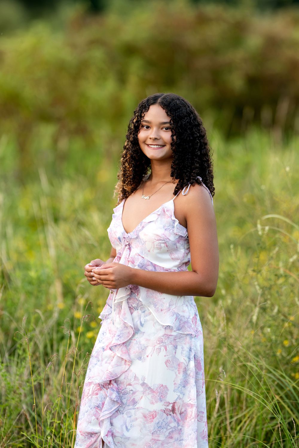 Smiling young woman in floral sundress standing in lush green field on summer day.