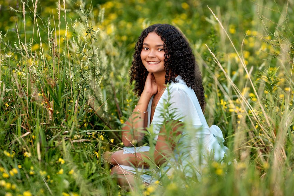 Young woman with curly hair smiling in a lush green meadow with yellow wildflowers on a sunny day.