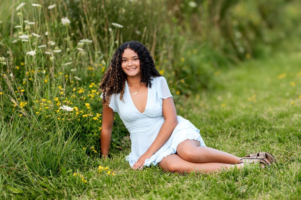 Young woman in white dress sitting on grass among wildflowers smiling outdoors.