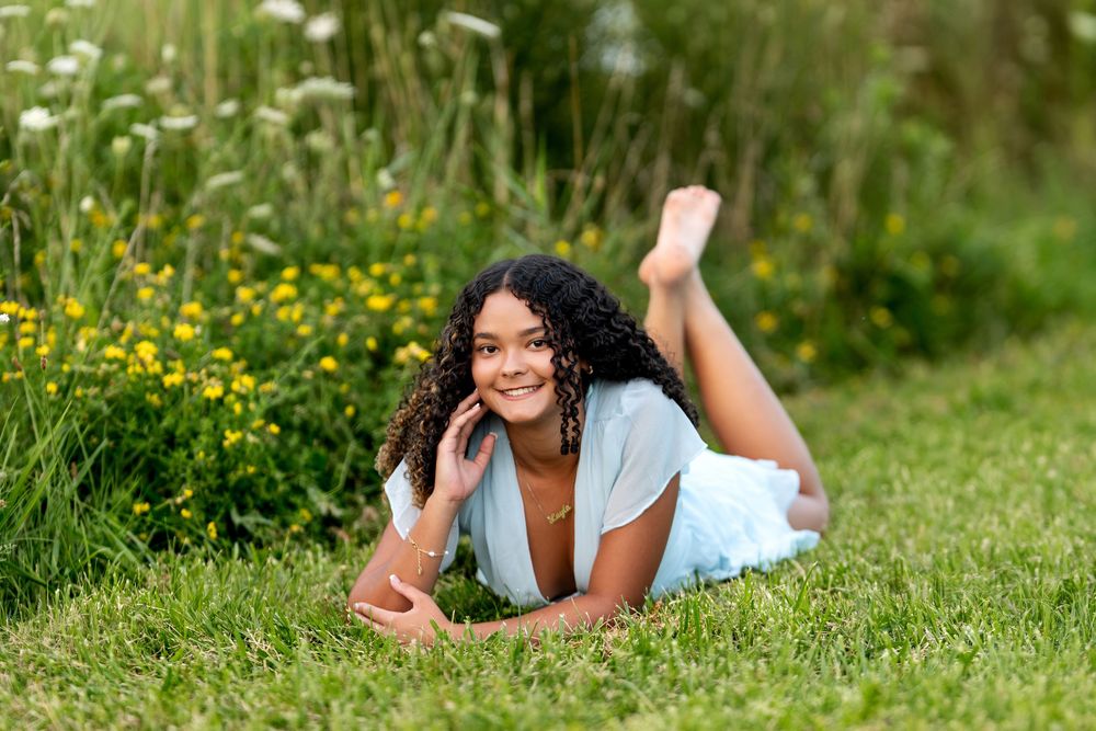 Happy young woman lying on green grass near yellow wildflowers in a light blue dress.