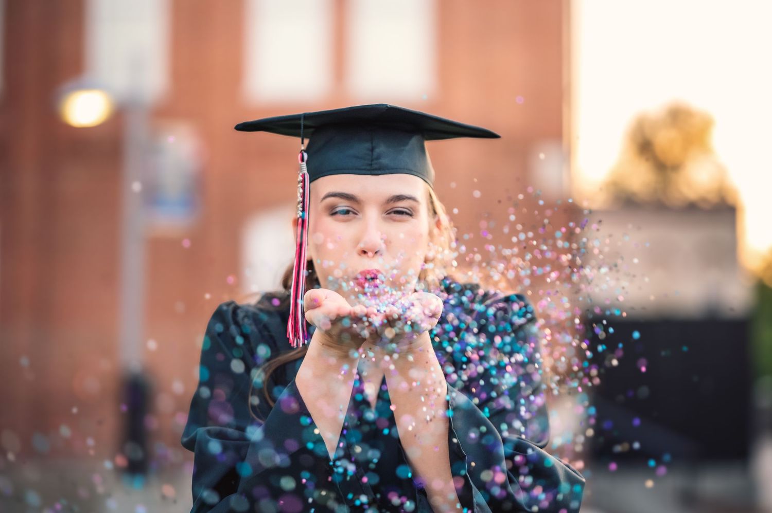 A female graduate in cap and gown blows colorful confetti from her hands outdoors at a university campus.