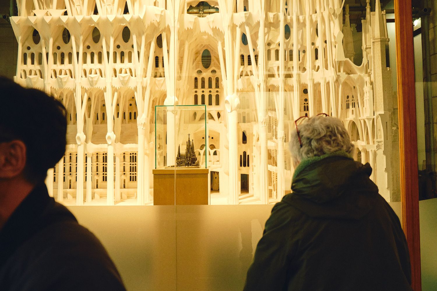 Two visitors admire an illuminated architectural scale model of the Sagrada Familia cathedral on display indoors.
