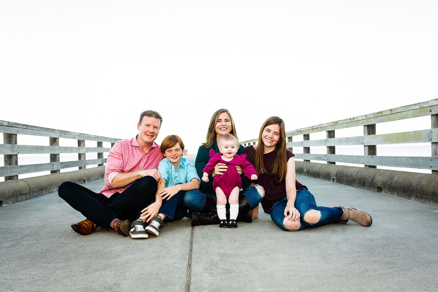 Happy family of five sitting together on a wooden pier bridge, smiling outdoors in casual colorful clothing.