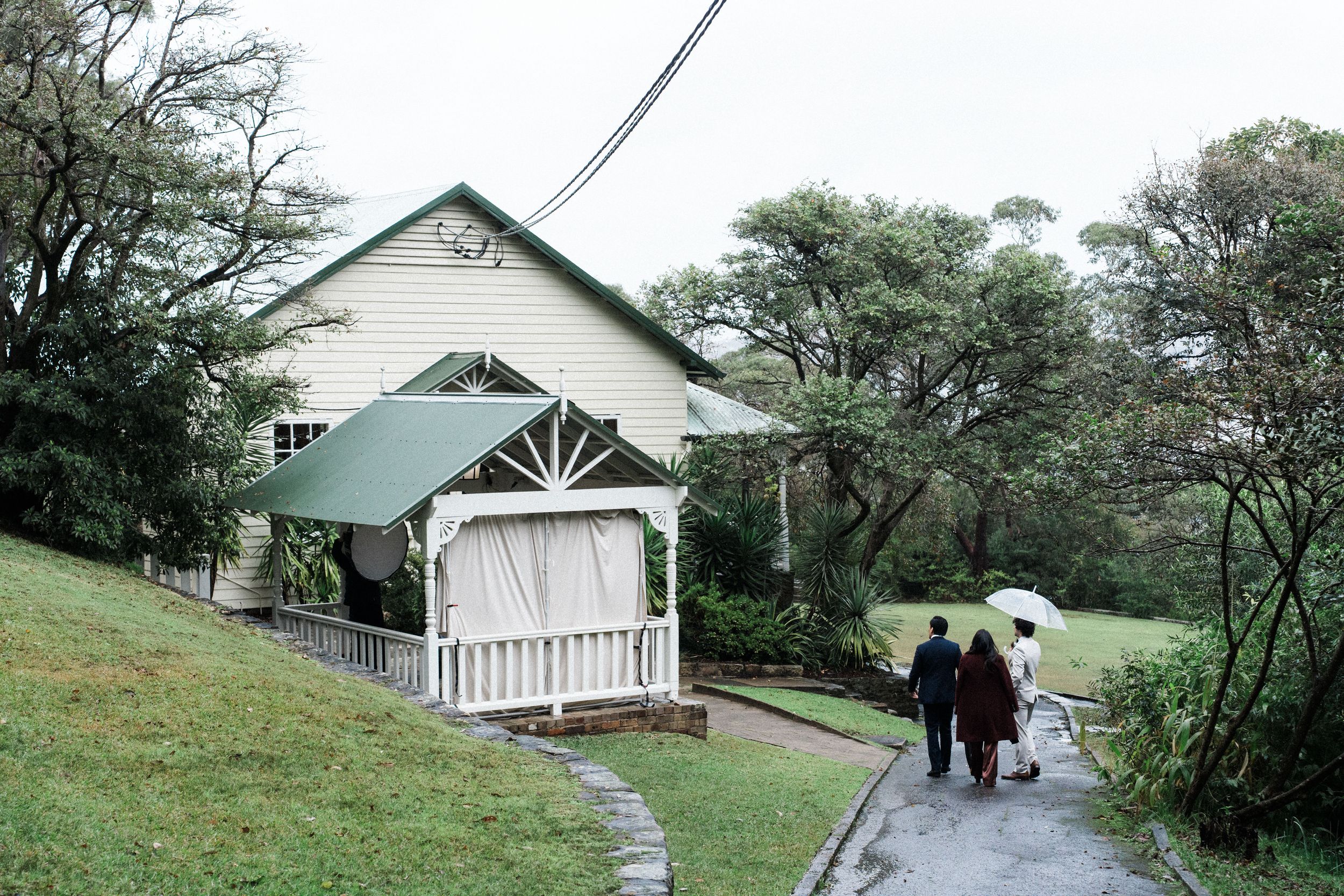 Guests walk along a path toward a charming white chapel with green roof surrounded by lush trees on a rainy day.
