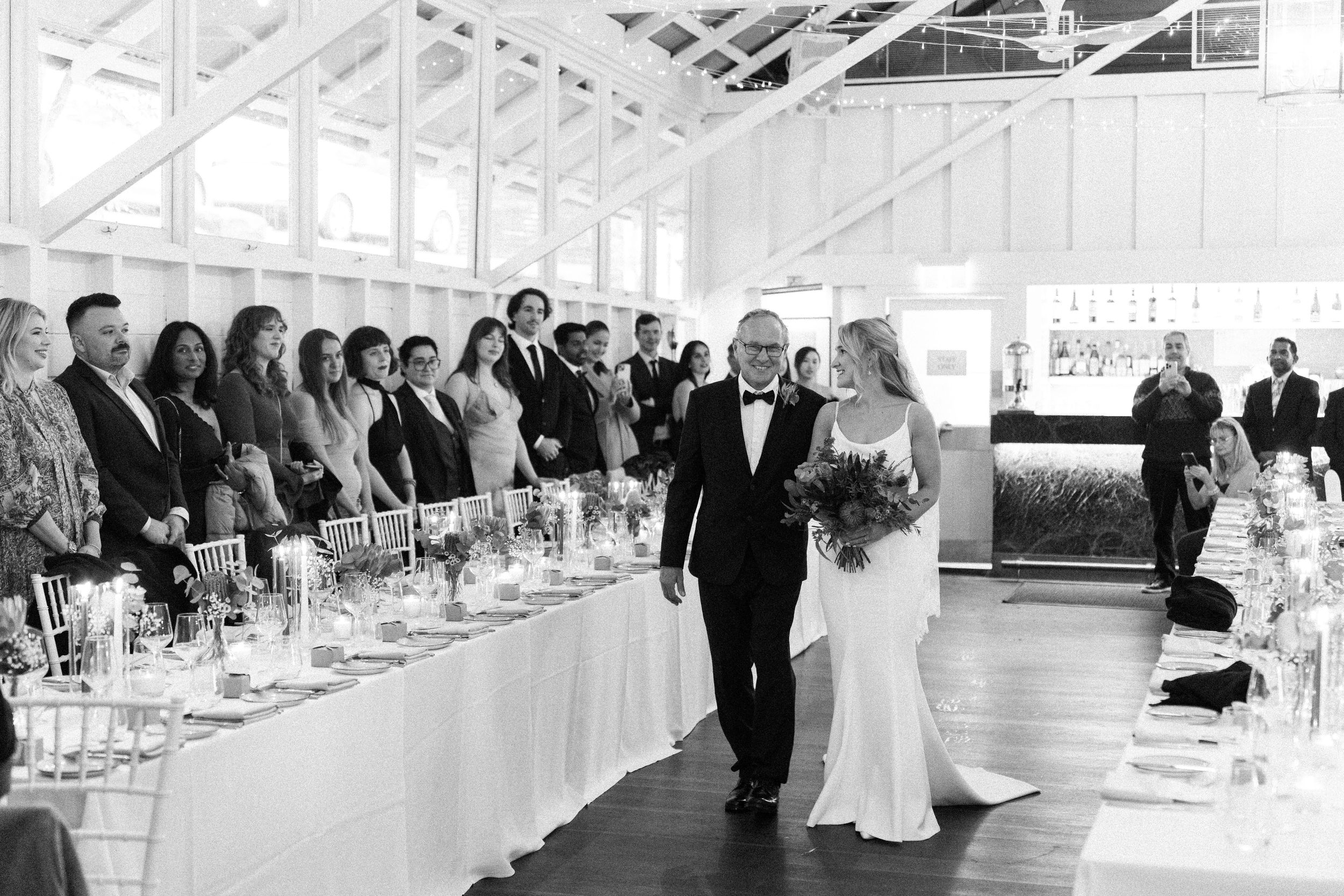 Black and white photo of a bride walking down the aisle with her father at an elegant indoor wedding reception.
