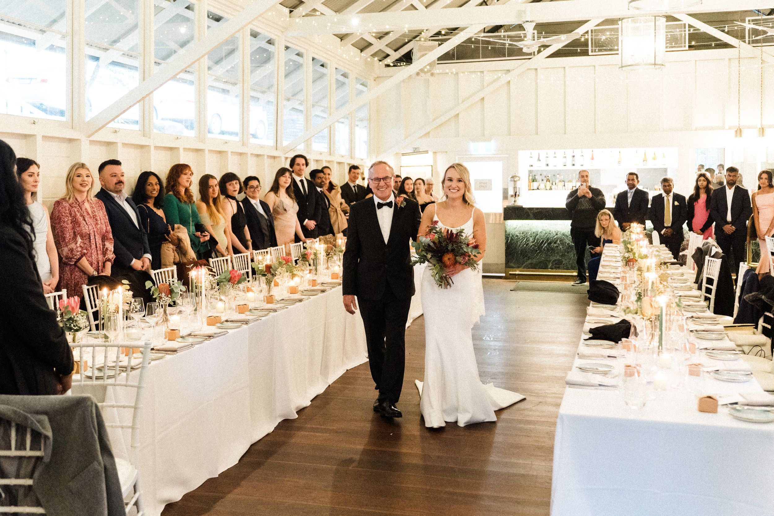 Bride walks down the aisle with her father in an elegant glass-walled wedding venue with gold table settings.