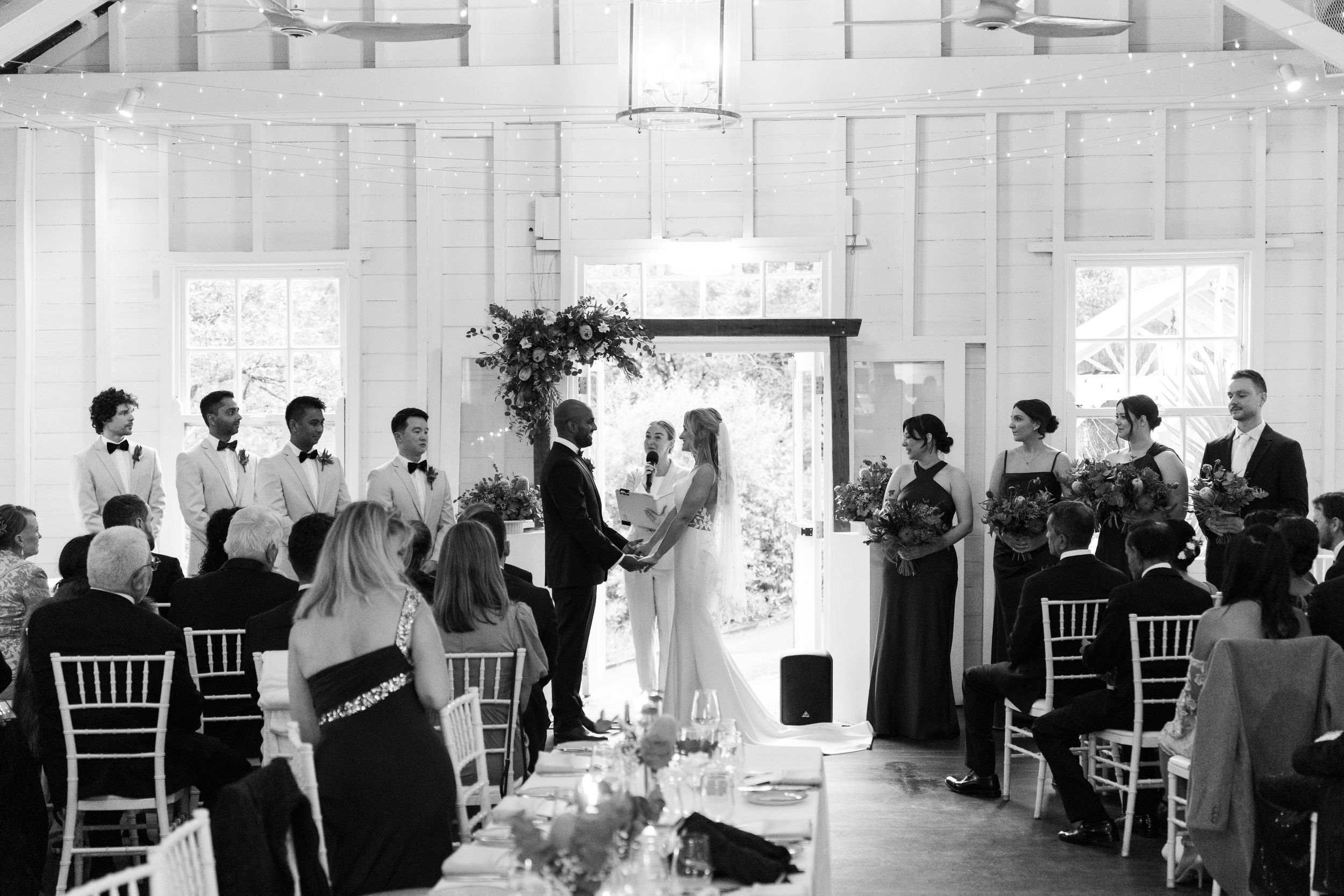 Black and white photo of an elegant indoor wedding ceremony with bride and groom at the altar surrounded by wedding party.