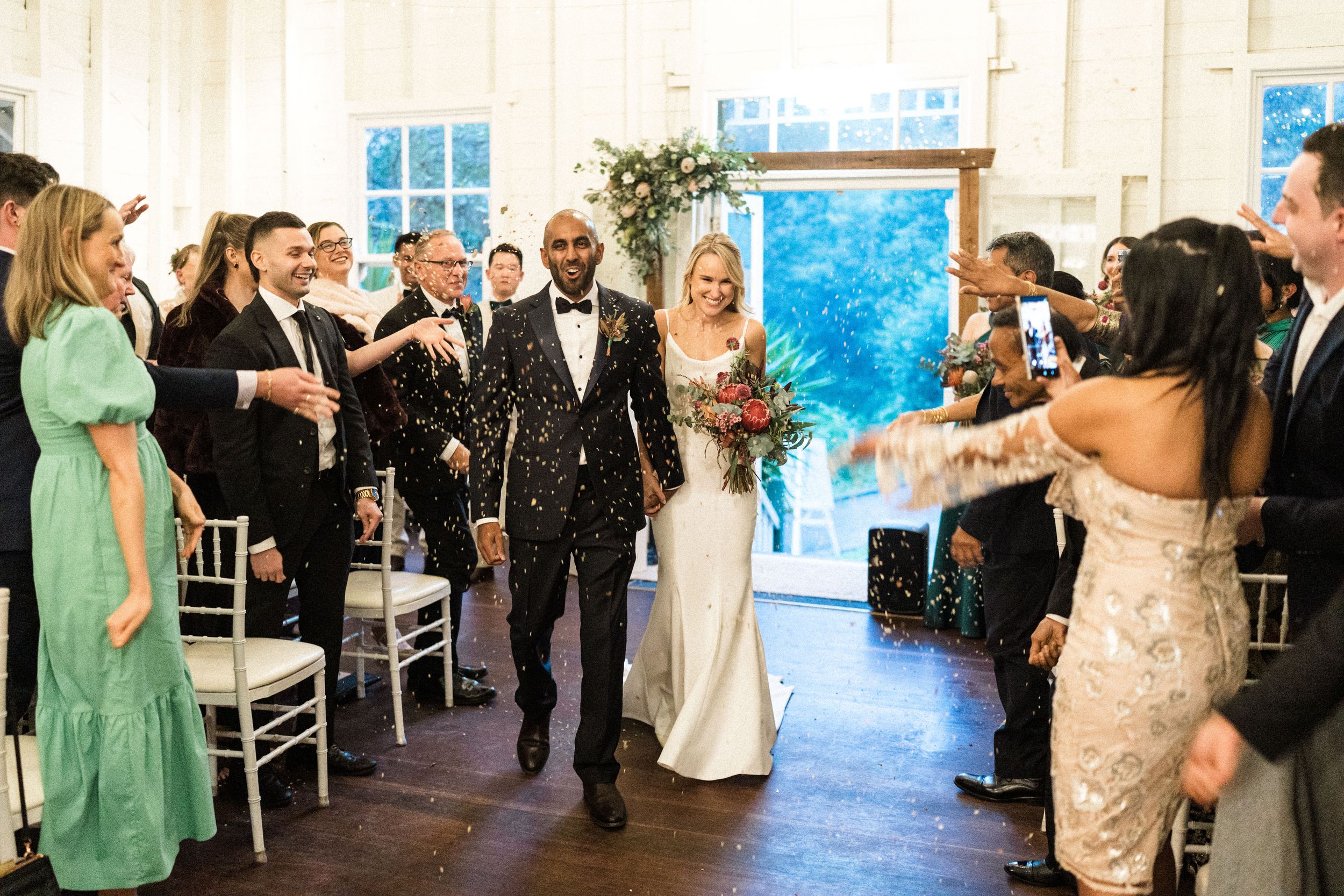 Newlywed couple walks down the aisle as guests celebrate with confetti in an elegant white wedding venue.