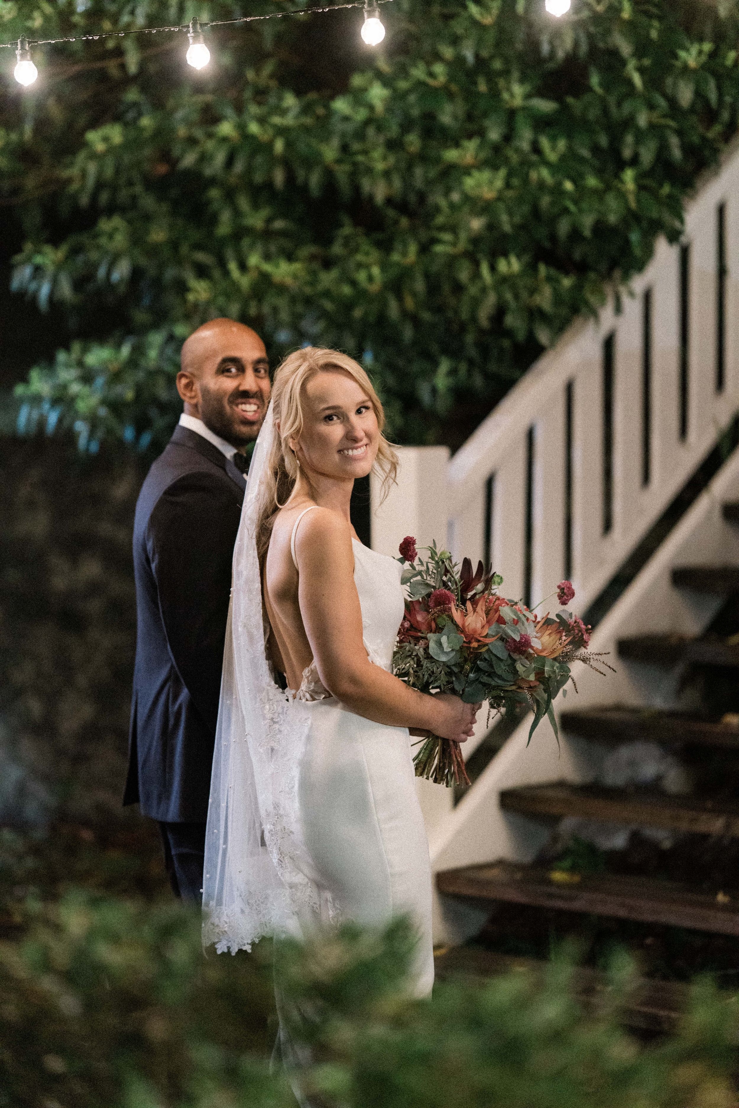 Bride in white gown holding bouquet stands with groom in navy suit near staircase under string lights at night.