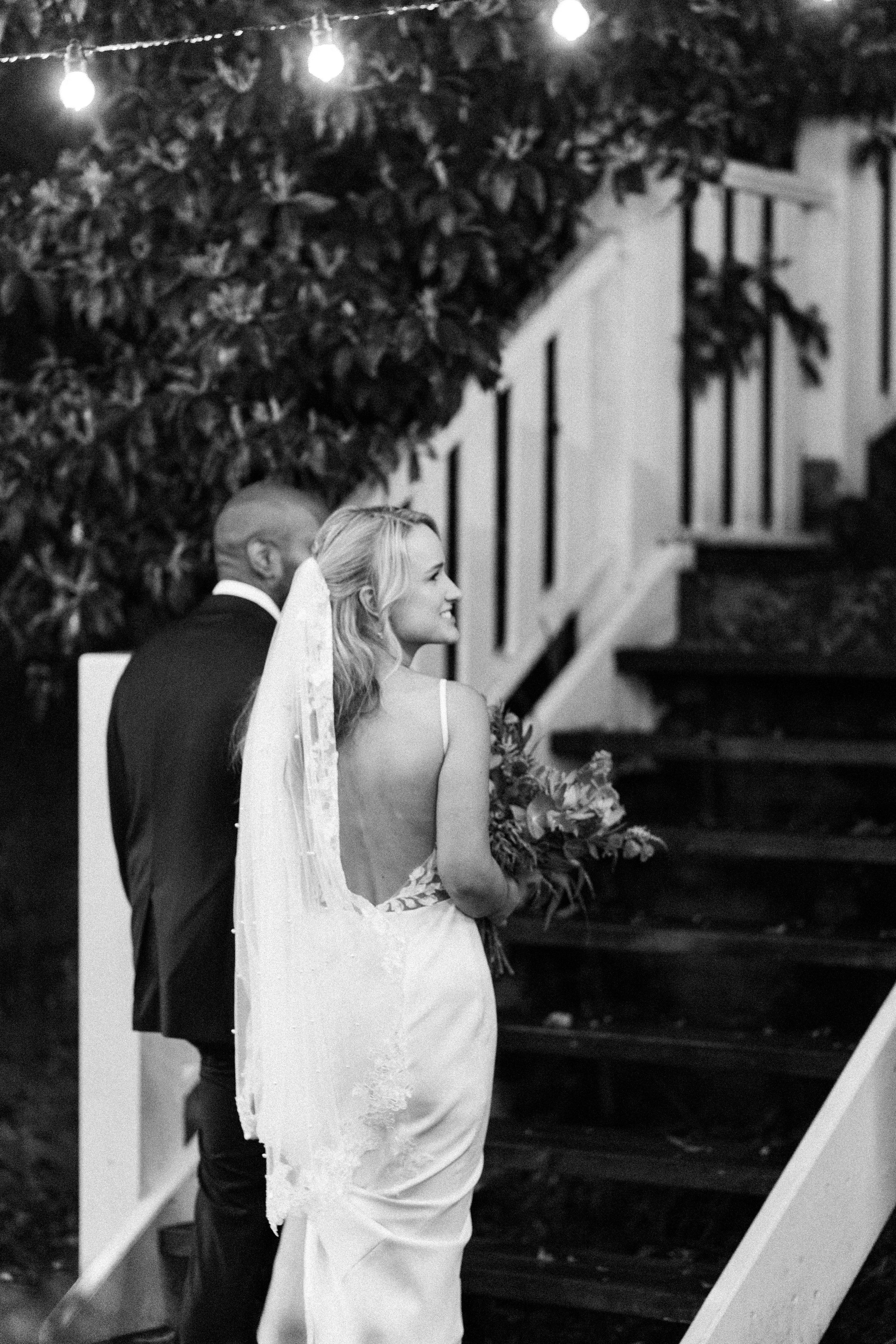 Black and white photo of a bride in a backless gown holding bouquet, smiling back at groom near staircase.