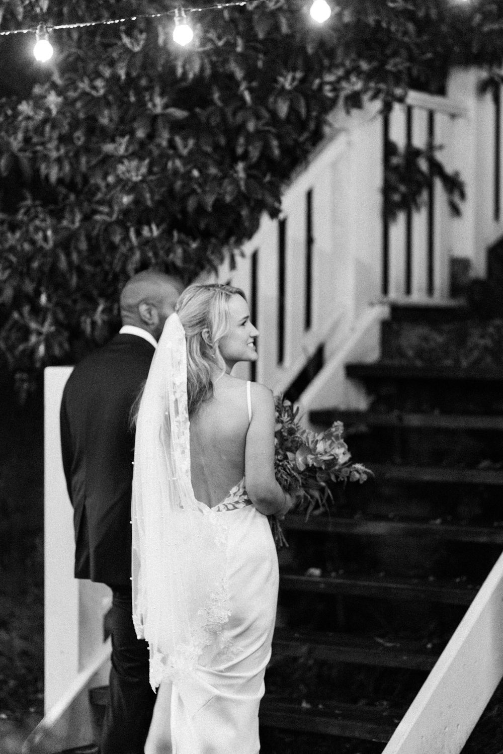 Black and white photo of a bride in a backless gown holding bouquet, smiling back at groom near staircase.