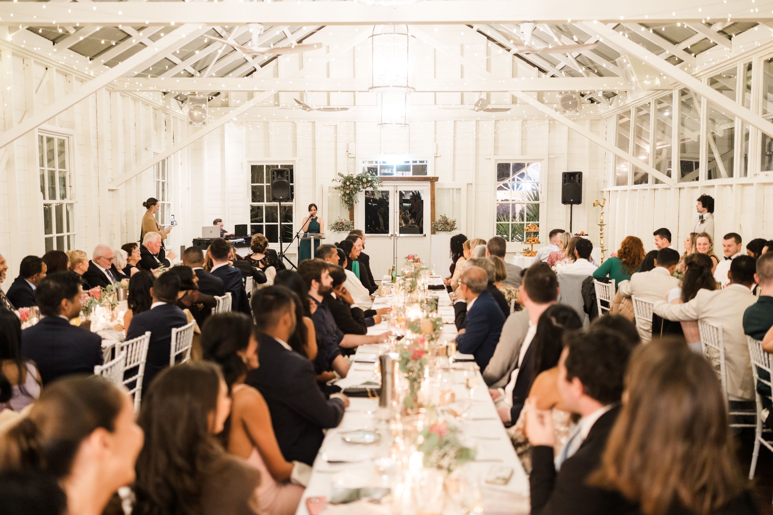 Elegant wedding reception in a white greenhouse venue with guests seated at long tables adorned with candles and floral centerpieces.