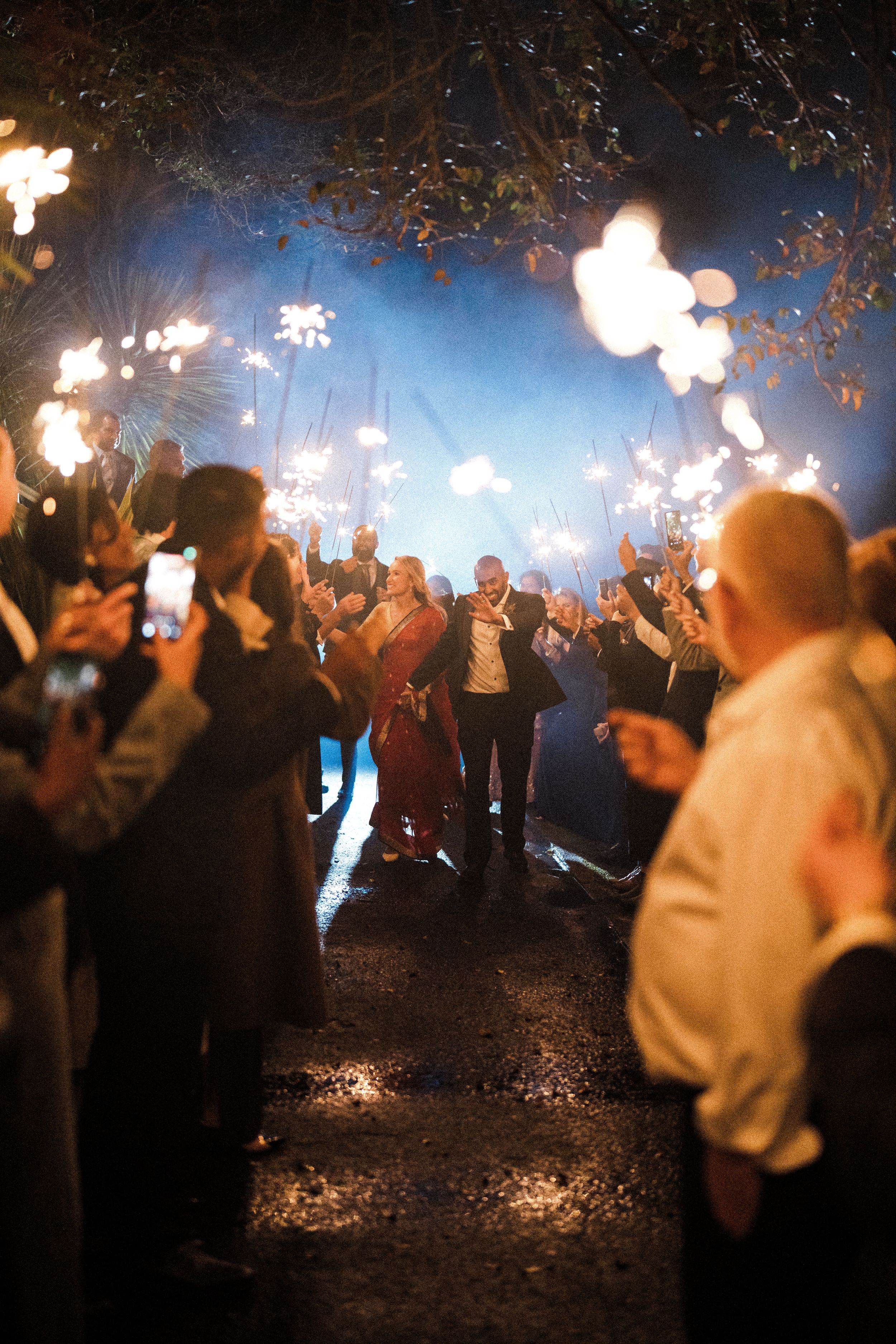 Newlyweds walk through a sparkler tunnel at night surrounded by cheering wedding guests holding sparklers.