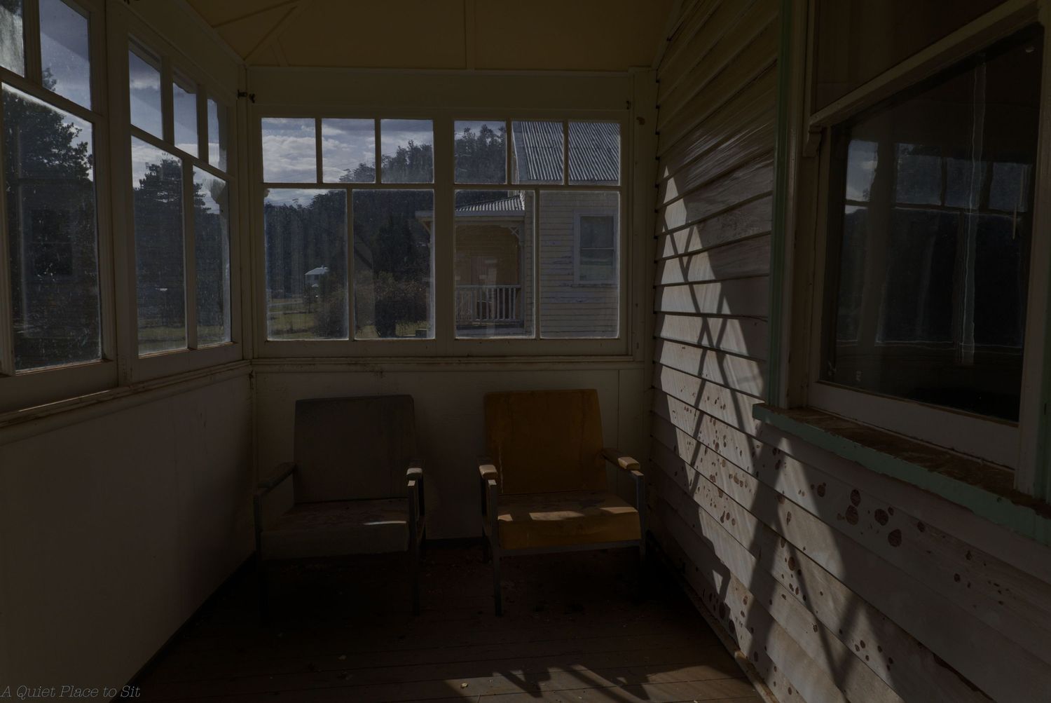 Two vintage armchairs sit in a quiet enclosed sunroom with white-framed windows and weathered wood siding, lit by soft shadows.