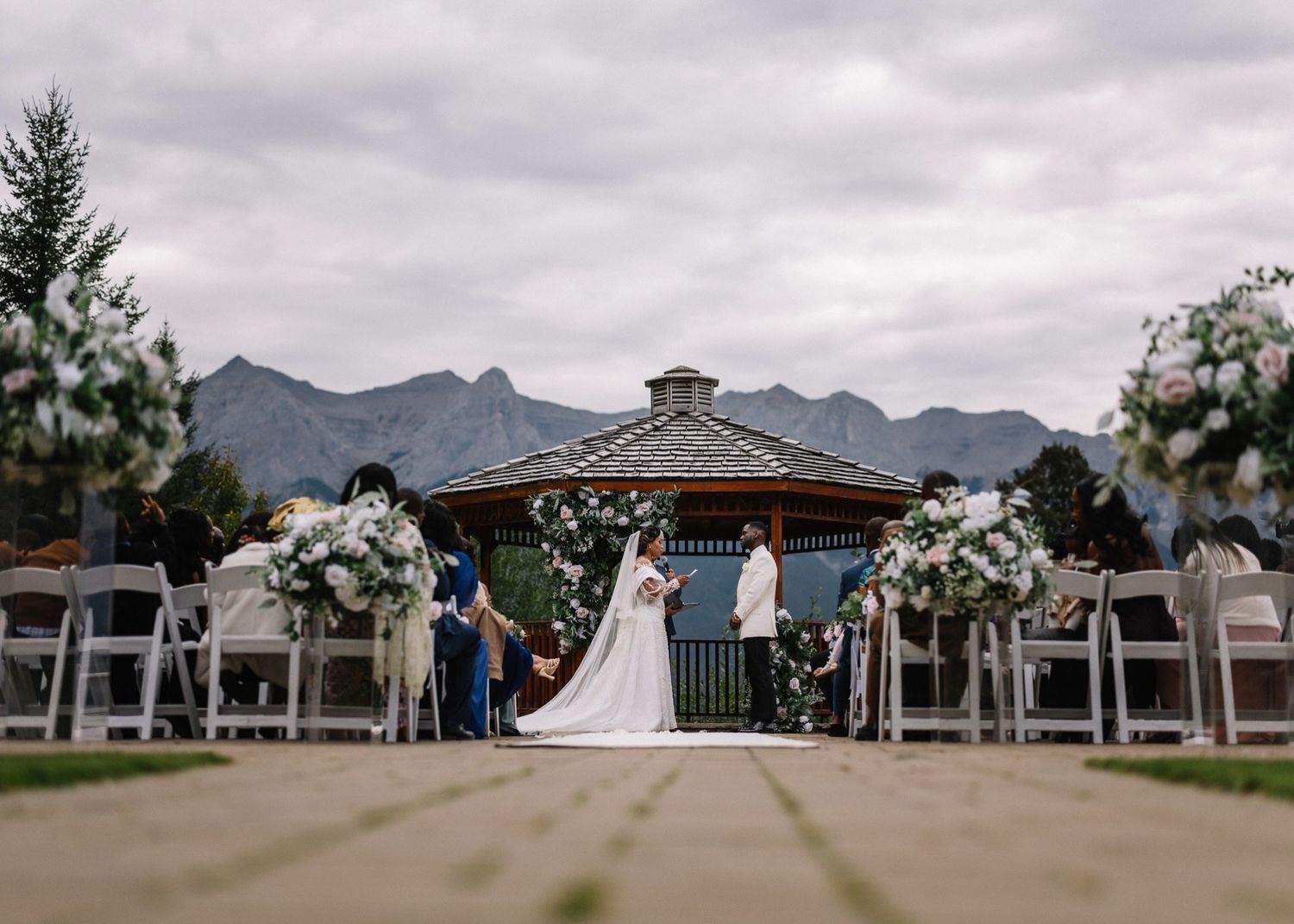 Bride and groom exchange vows at an outdoor mountain wedding ceremony under a gazebo with floral arrangements and guests seated nearby.