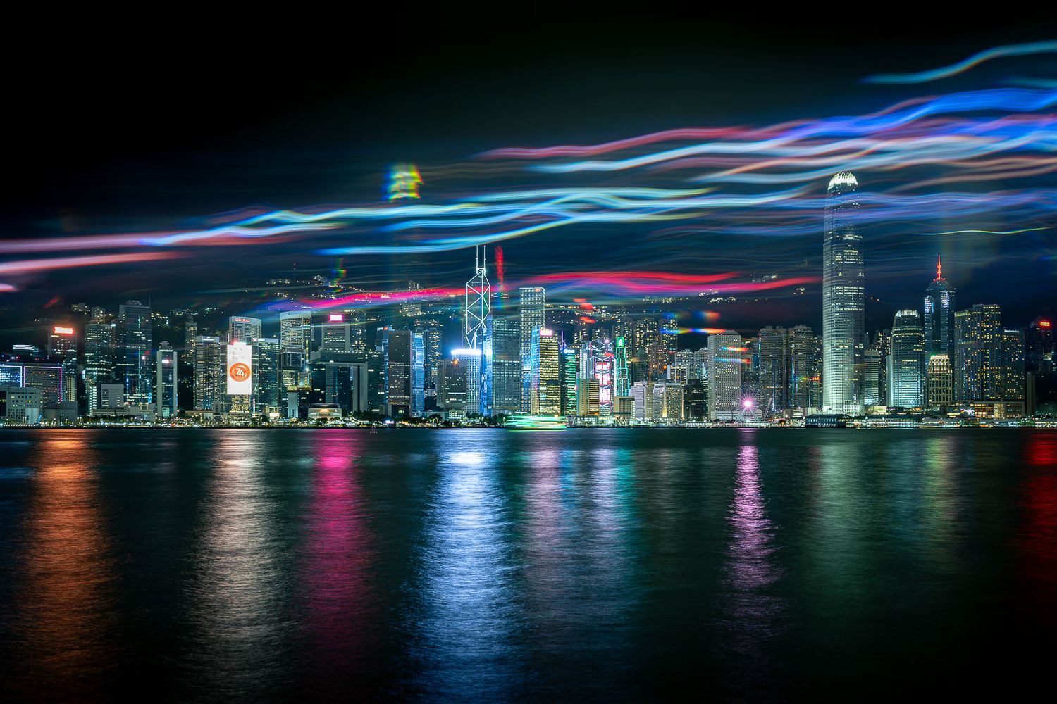 Colorful light trails streak across the night sky above a glowing city skyline reflected in the calm harbor waters.