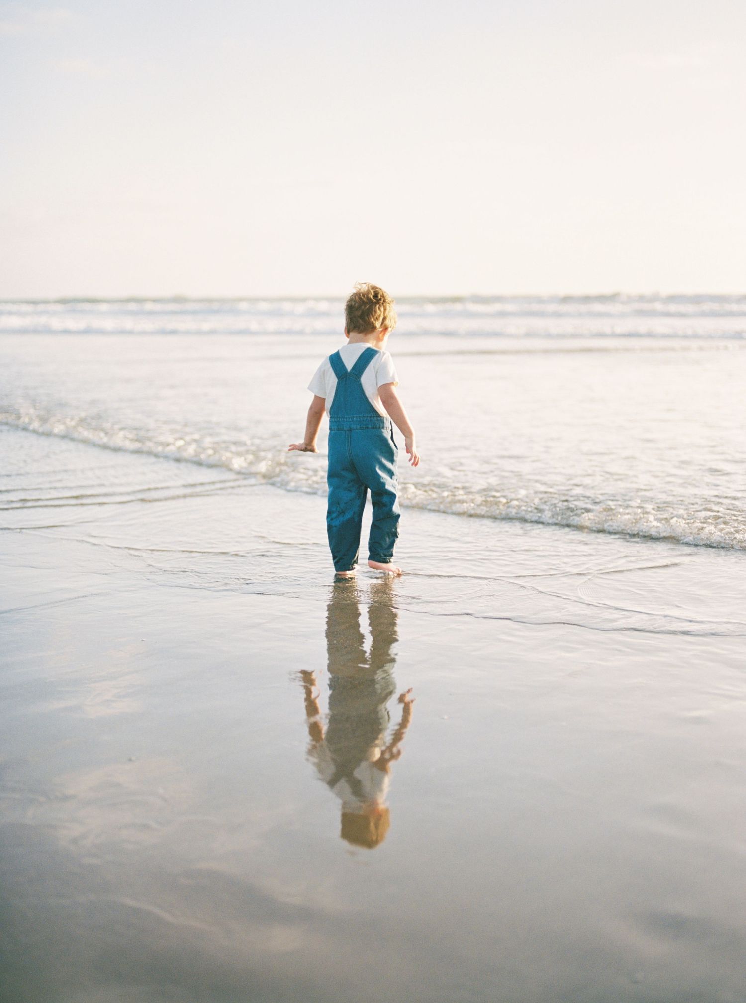 Toddler in blue denim overalls walking barefoot along the wet sandy beach shoreline at sunset with reflection in the water.