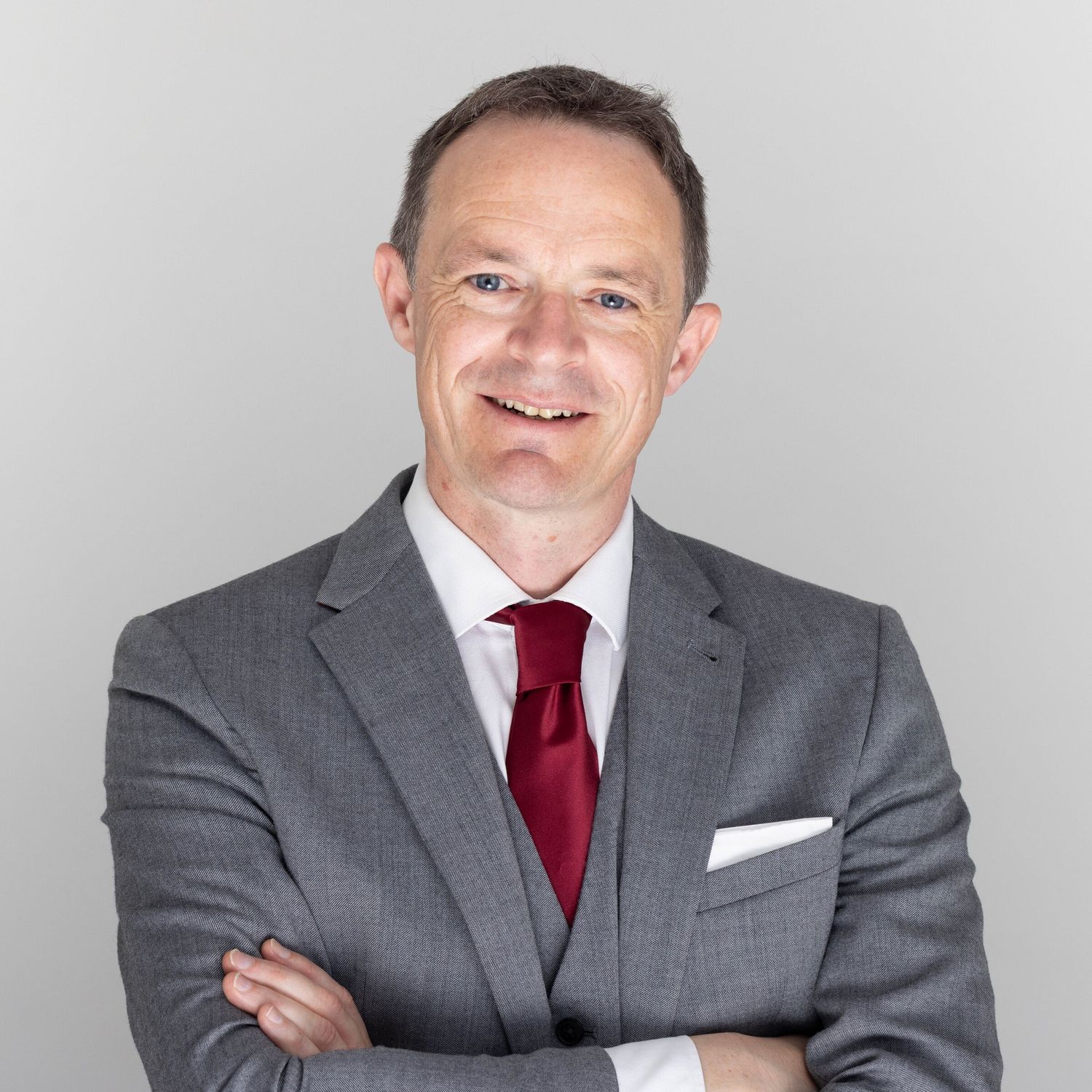 Corporate headshot of a smiling businessman in a grey three-piece suit and red tie on a pale grey background