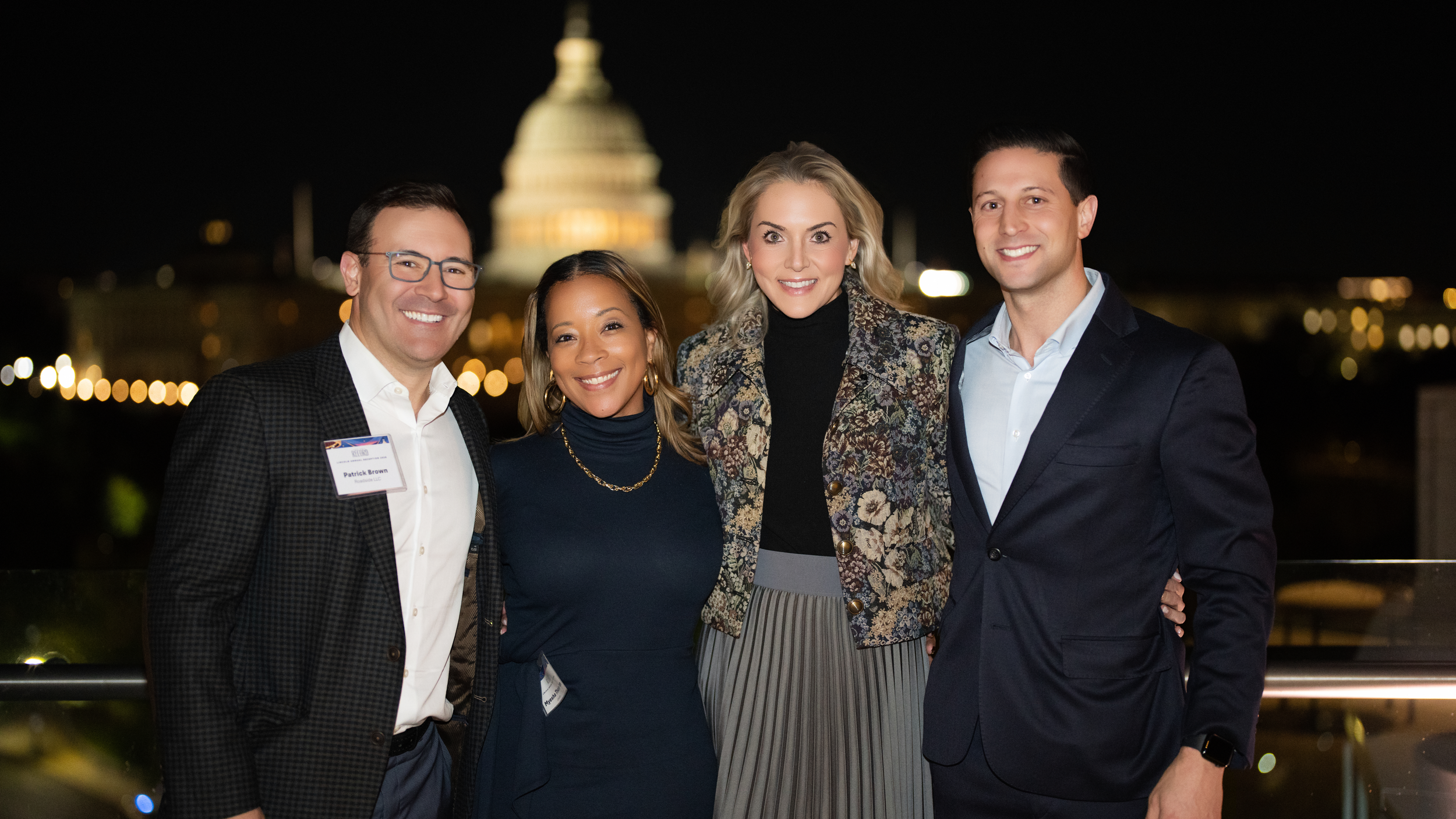 Four guests pose on the 101 Constitution rooftop at night with the illuminated U.S. Capitol behind them.