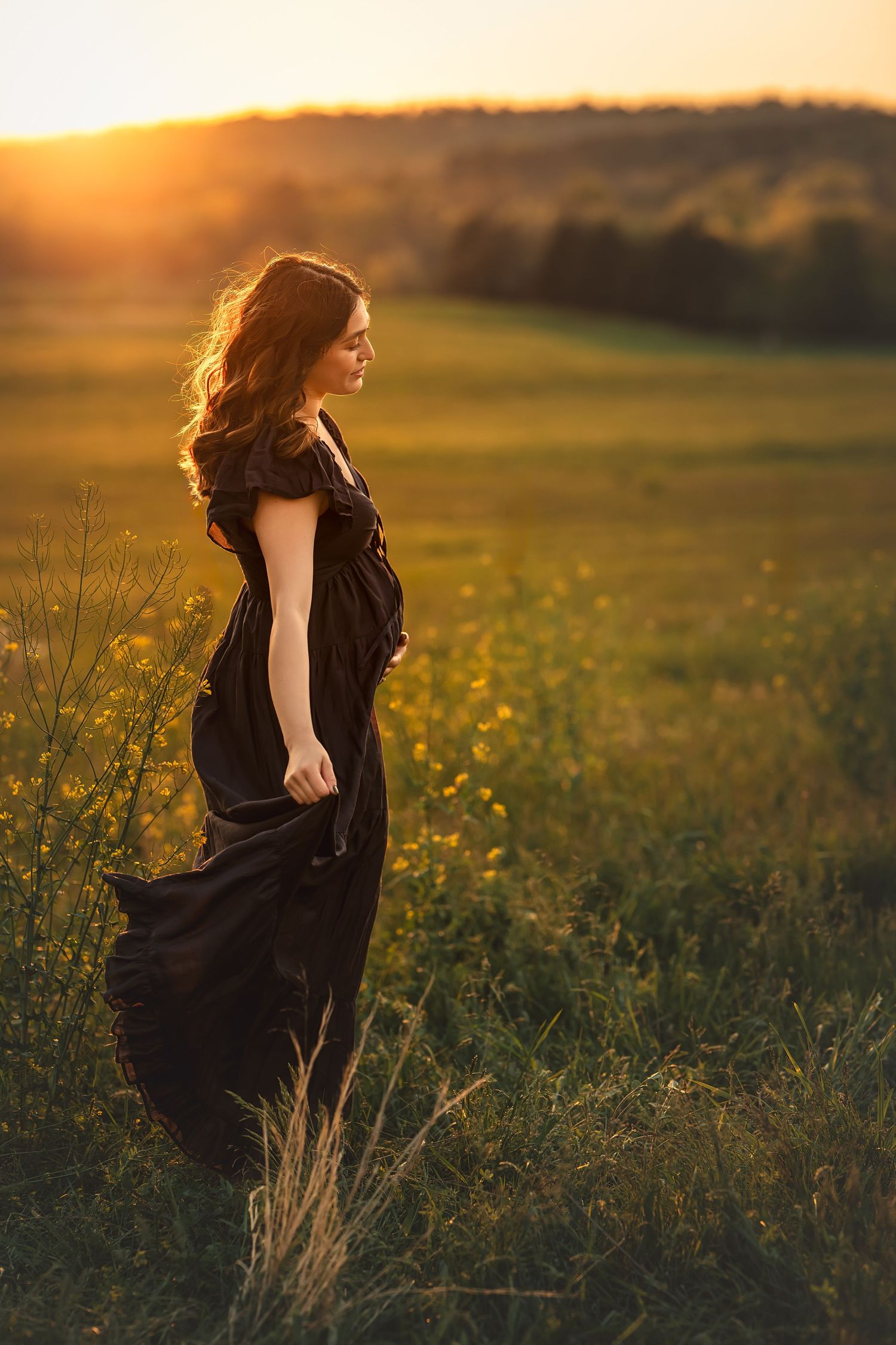 Woman in black dress standing in golden sunset field with wildflowers and rolling hills in background.